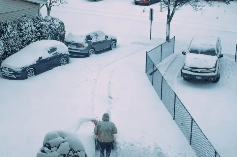 A buried street of snow-covered cars with one person shovelling alone at dawn