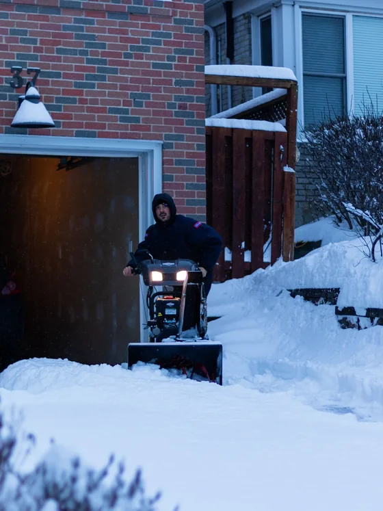 An operator running a snow blower up a residential driveway before dawn