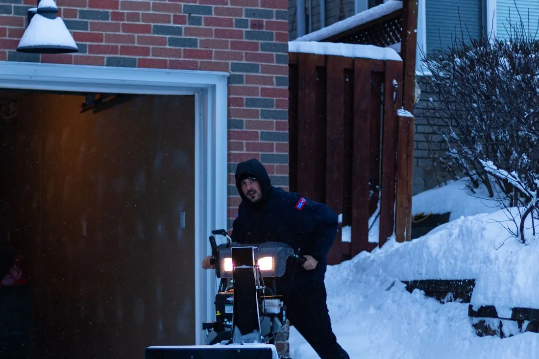 An operator starting a snow blower under headlights beside a garage